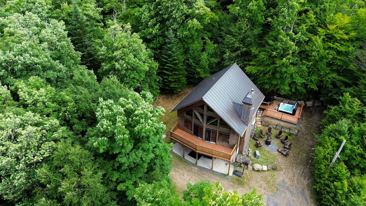 A cozy chalet is surrounded by lush green trees, providing a serene and secluded atmosphere. The structure features a spacious deck and is complemented by a hot tub positioned near an outdoor seating area with several chairs, creating an inviting space for relaxation.