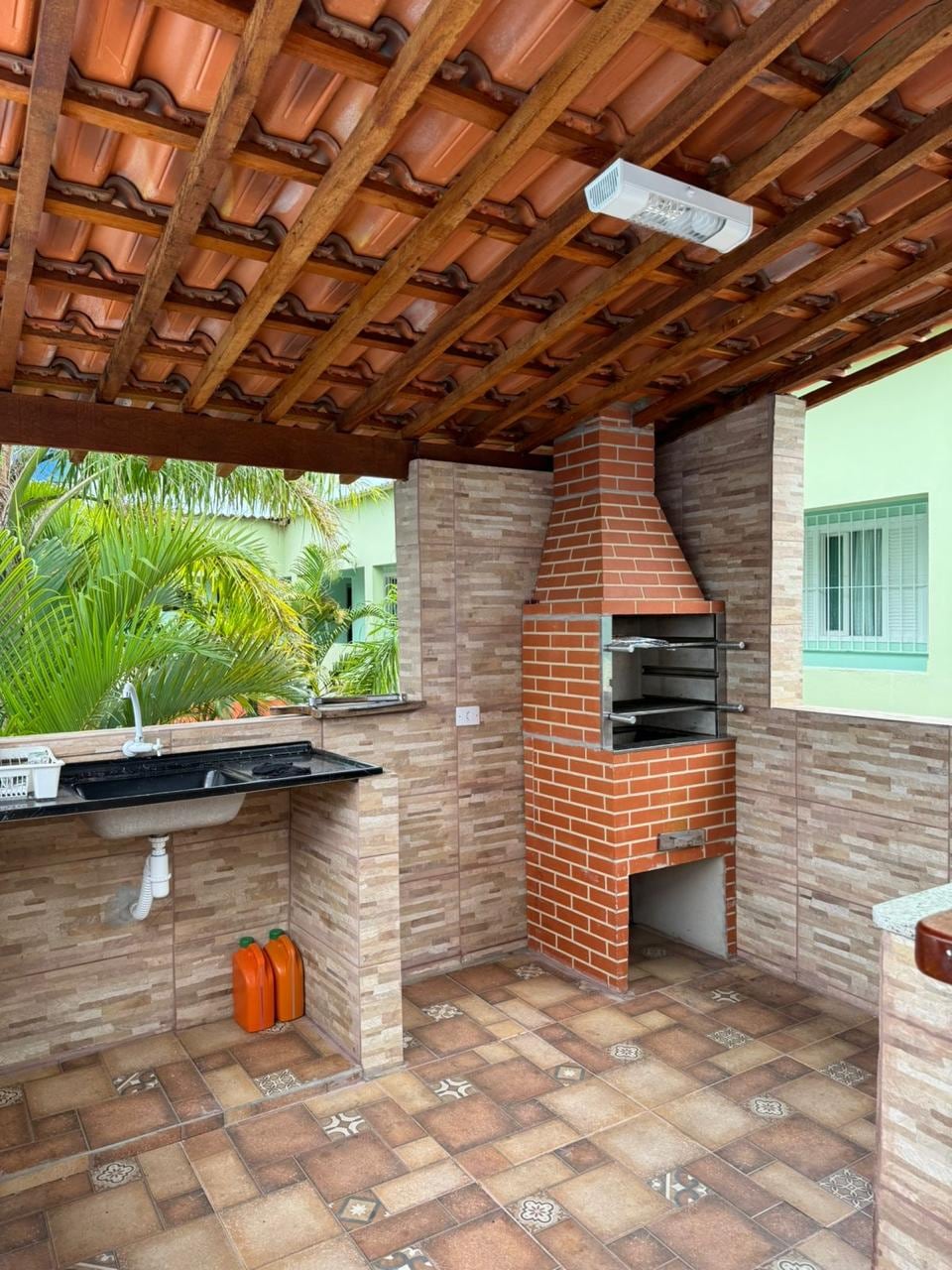 An outdoor barbecue area is featured with a brick grill and a stone countertop. The surrounding walls are decorated with patterned tiles. Green foliage adds a natural touch to the inviting space, while appliances and storage can be seen in the work area.
