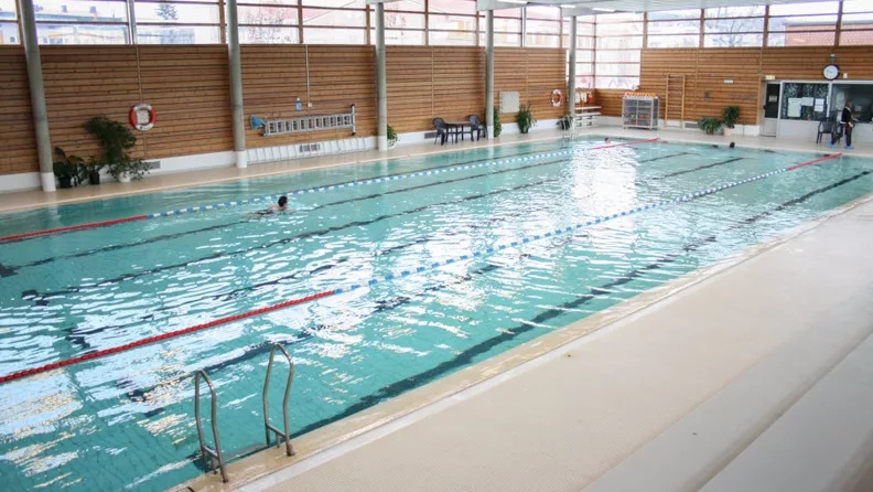 An indoor swimming pool is visible, featuring clear, blue water with marked lanes. Sunlight filters in through large windows, illuminating the area. Lounge chairs are arranged alongside the pool, and safety equipment is mounted on the walls. A person is seen swimming in the distance.