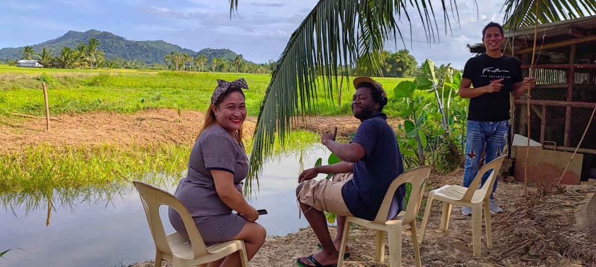 Two individuals are seated on light-colored plastic chairs near a tranquil pond. Lush green fields stretch into the background, framed by distant hills. A palm tree provides shade, adding to the serene atmosphere of the setting.