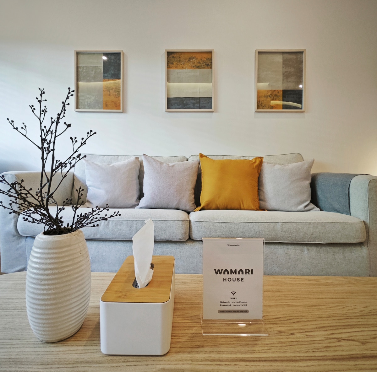 A modern living room features a three-seater sofa adorned with neutral and yellow cushions. A stylish vase with dark branches sits atop a wide coffee table. Artwork in light frames decorates the wall behind, contributing to the room’s contemporary aesthetic.