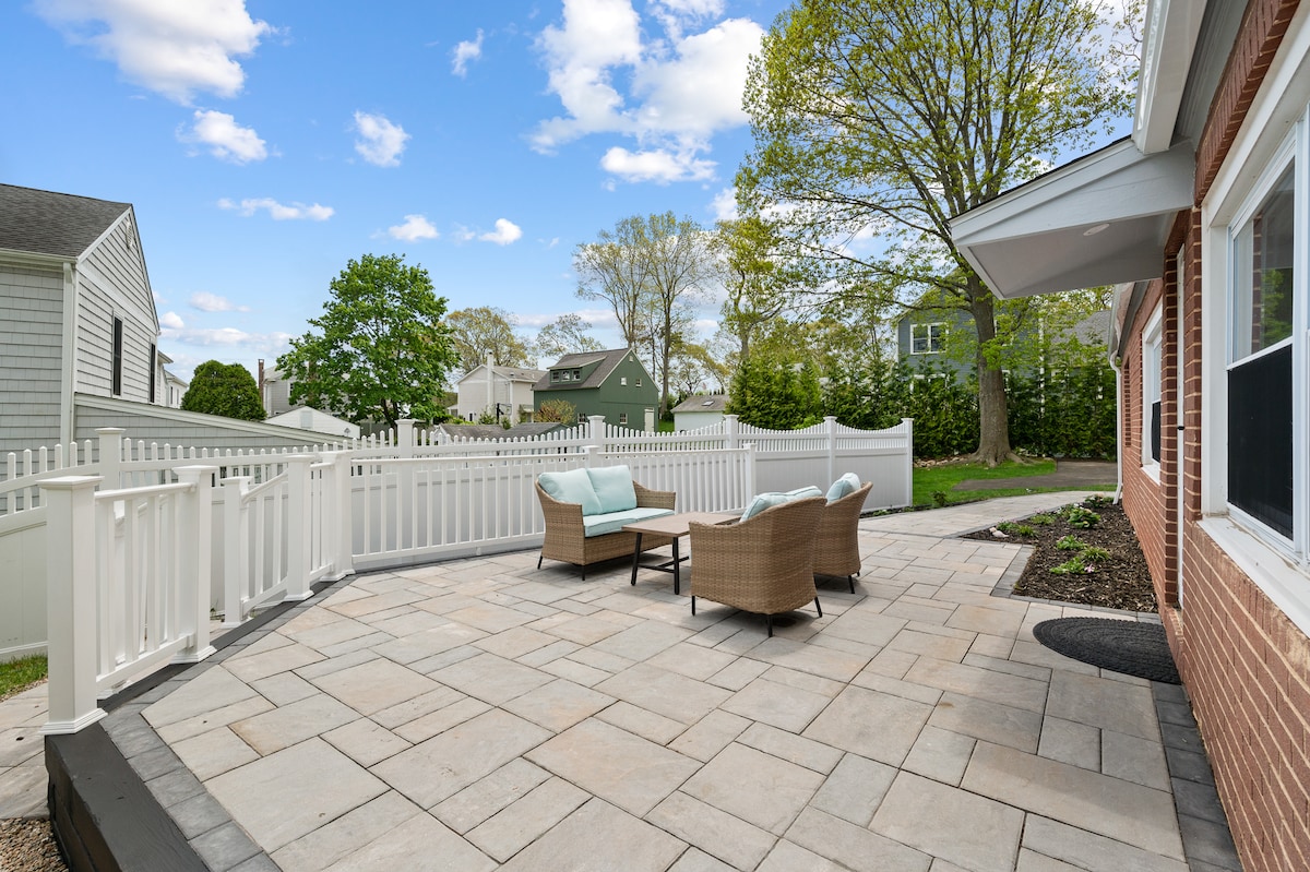 A spacious outdoor patio is depicted, featuring a set of light blue cushions on a wicker seating arrangement. A stone surface provides a neutral base, while a white picket fence encloses the area, creating an inviting space surrounded by greenery and trees.