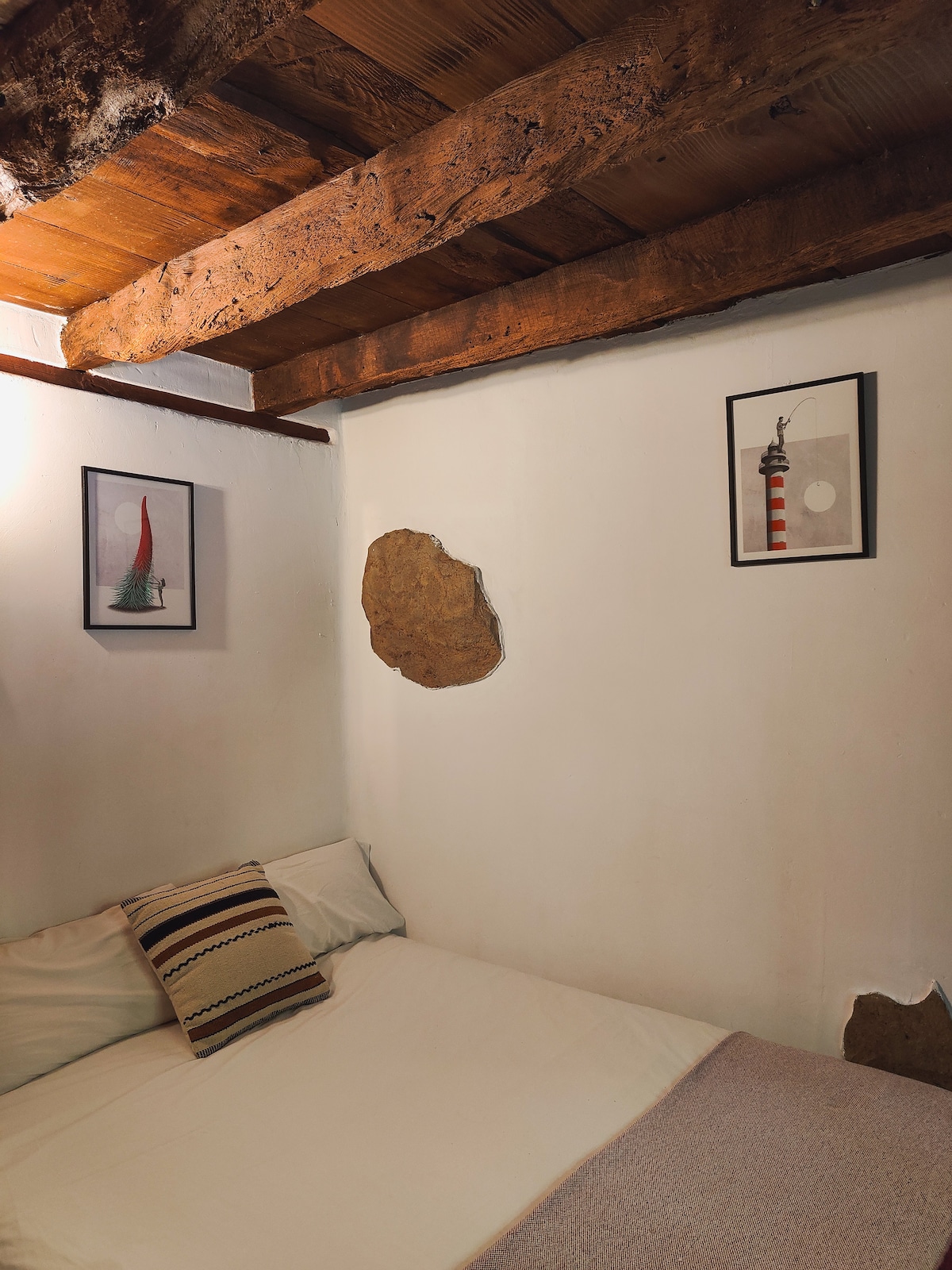 A cozy bedroom features exposed wooden beams and textured walls with natural stone accents. Two framed artworks adorn the walls above a neatly made bed, which includes a colorful striped pillow. Soft natural light brightens the space, creating a restful environment.
