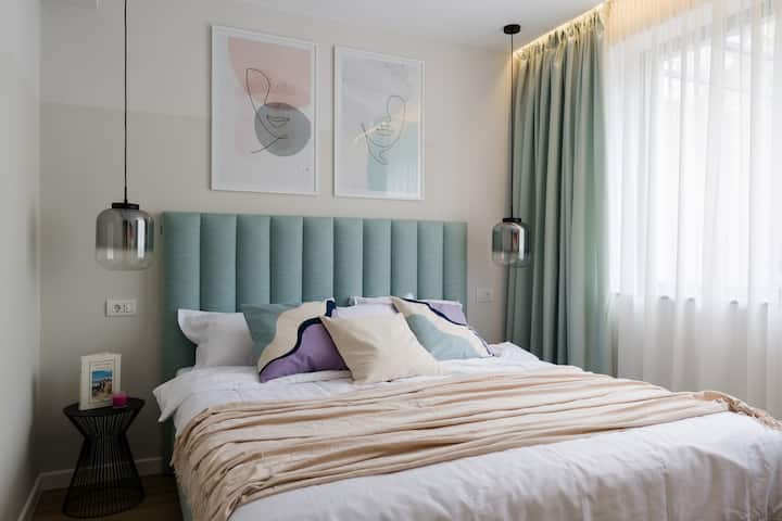 חדר שינה מואר ומזמין עם ראש מיטה מרופד בצבע ירוק מנטה, כריות בשכבות, שמיכה בצבע בז' ווילונות באורך מלא. החדר מלא באור טבעי שמייצר אווירה רגועה ונינוחה.