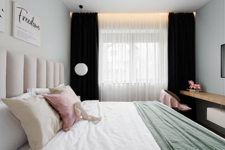 חדר שינה מודרני עם גווני פסטל עדינים, וילונות האפלה מלאים וחלון גדול לאור טבעי. נוח ואלגנטי, מושלם ללילות מרגיעים ולבוקרים מסוגננים.