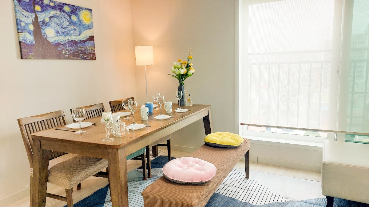 A dining area is set with a wooden table and four chairs, surrounded by a light rug. Fresh flowers are displayed in a vase, while colorful cushions sit on the benches nearby. Soft lighting from a lamp enhances the inviting atmosphere.