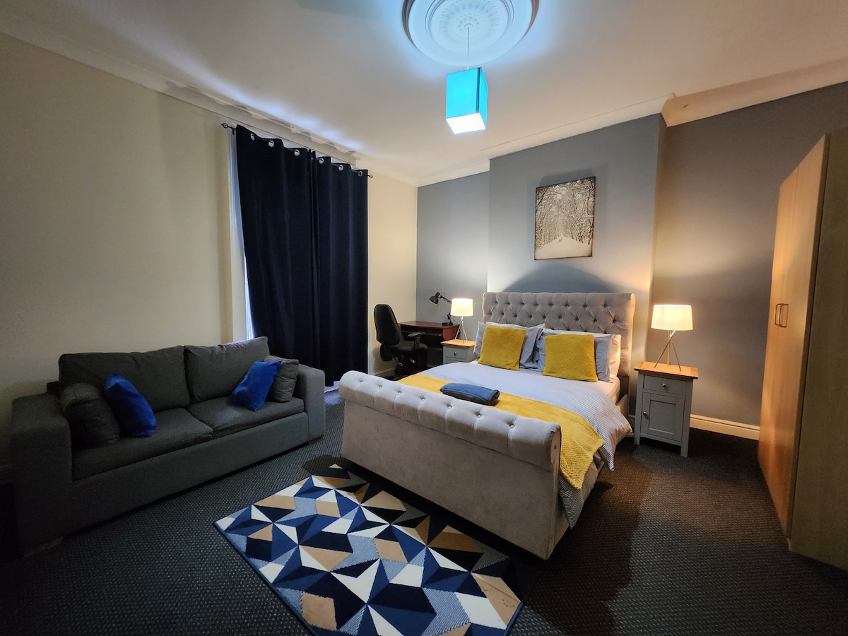 A spacious bedroom features a large bed with yellow and blue accents. A comfortable sofa is situated nearby, complemented by a patterned area rug. Two bedside lamps provide soft lighting, while a desk and chair are positioned against the wall. Dark curtains frame the window.