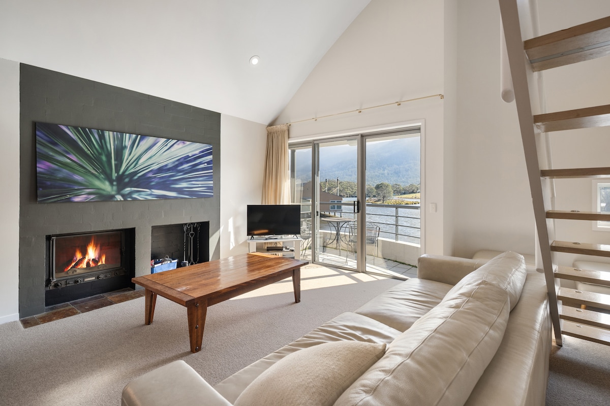 The living area features a comfortable lounge with a light-colored leather sofa, a wooden coffee table, and a large wall-mounted TV. A glass door opens onto a lakeside balcony, while a glass-fronted fireplace adds warmth to the serene space overlooking the scenic views.