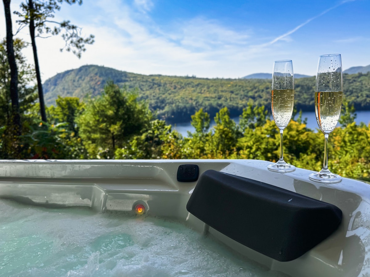 A private hot tub is situated outdoors, surrounded by greenery and offering views of the lake and mountains in the background. Two glasses of sparkling wine are placed on the tub's edge, reflecting the natural scenery under a clear blue sky.
