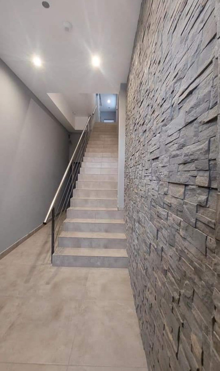 A modern staircase is set against a textured stone wall, with soft lighting illuminating the steps. The neutral color palette of the walls and floor contributes to a calming environment, while the railing provides a safe transition to the upper level.