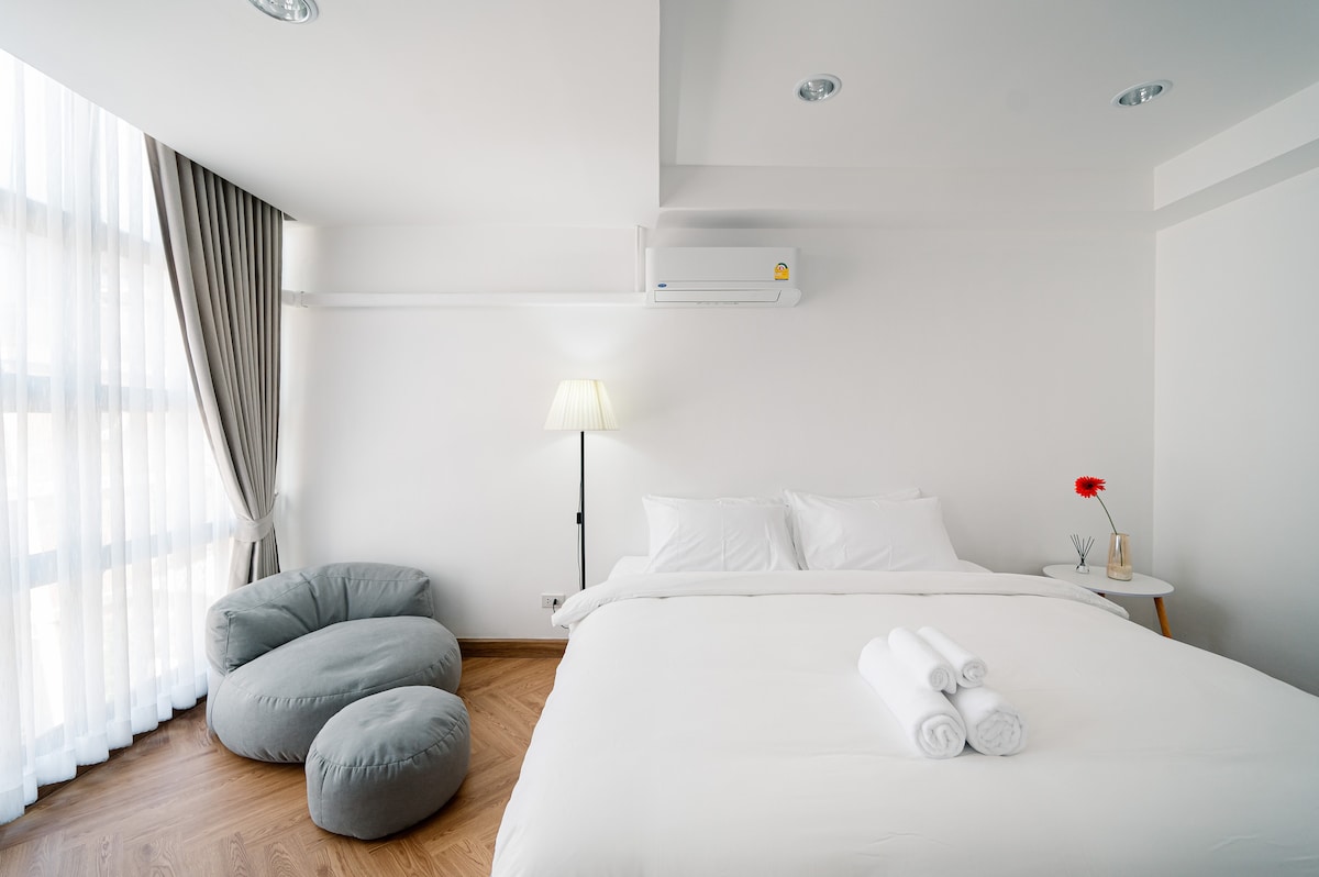 A well-lit room features a spacious bed with layered white bedding. A sleek bedside table holds decorative items. A floor lamp provides soft illumination, while a cozy bean bag chair adds a relaxed seating option. Large windows draped in sheer curtains allow natural light to filter in.
