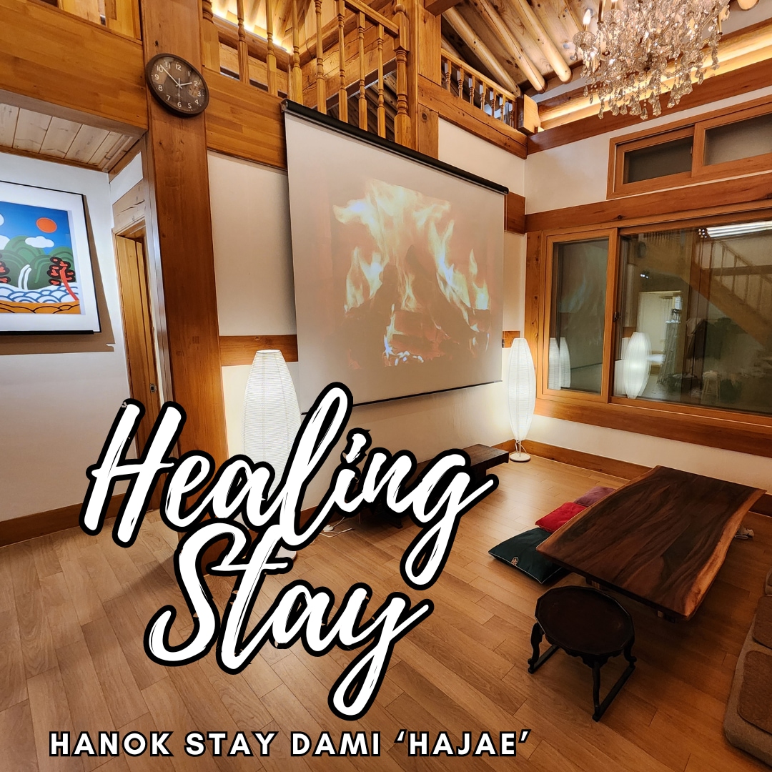 A welcoming indoor space features a large projection screen displaying a calming fireplace scene. Natural wood finishes and soft lighting from floor lamps create a serene environment. A low wooden table sits on the floor, enhancing the cozy atmosphere.