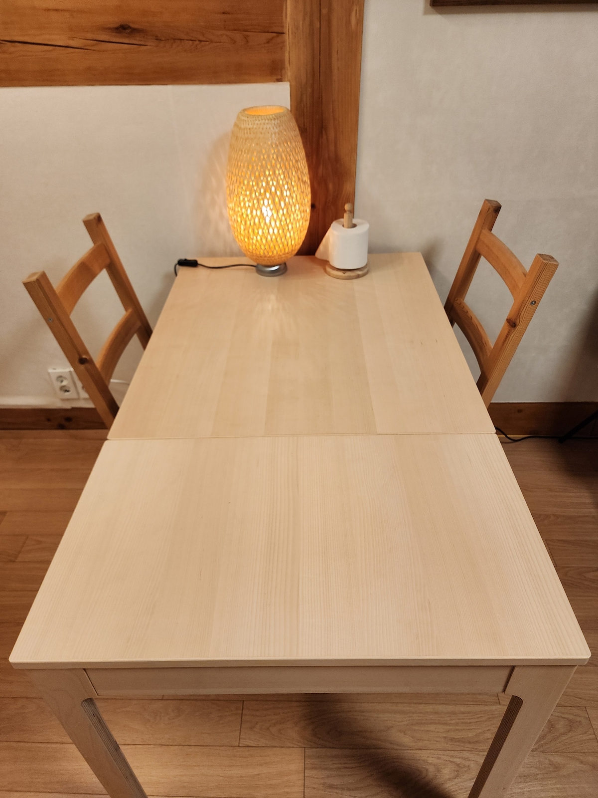 A light wooden table is shown with two simple wooden chairs positioned at either side. An ambient light fixture with a woven design is placed on the table, providing a soft glow. The flooring is comprised of warm-toned wood.
