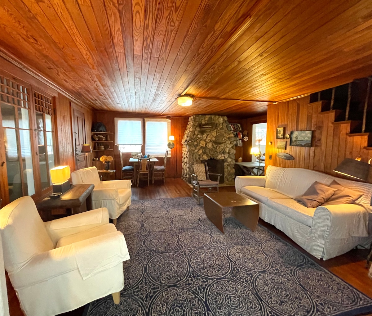 The living area showcases original hardwood floors and a river rock fireplace, surrounded by soft seating arranged on an area rug. Natural light filters in through the windows, highlighting a dining table in the adjacent space.