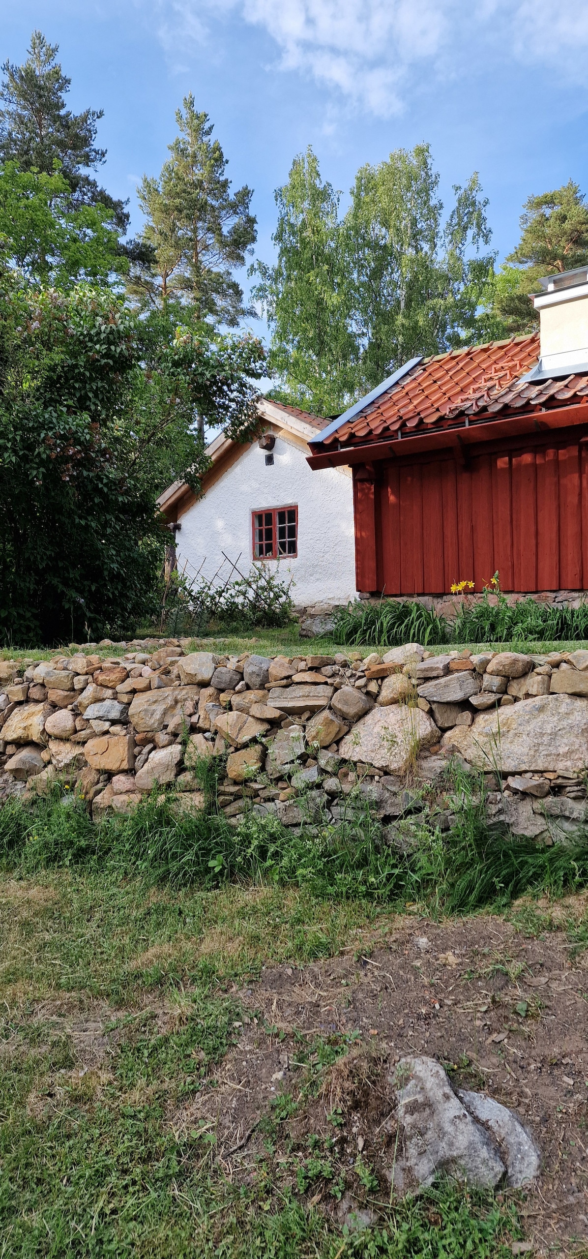 The image features a charming cottage positioned beside a natural stone wall. Lush greenery and trees form the backdrop, while a vibrant garden in front adds color. The cottage displays a combination of white and red walls topped with a traditional tiled roof.