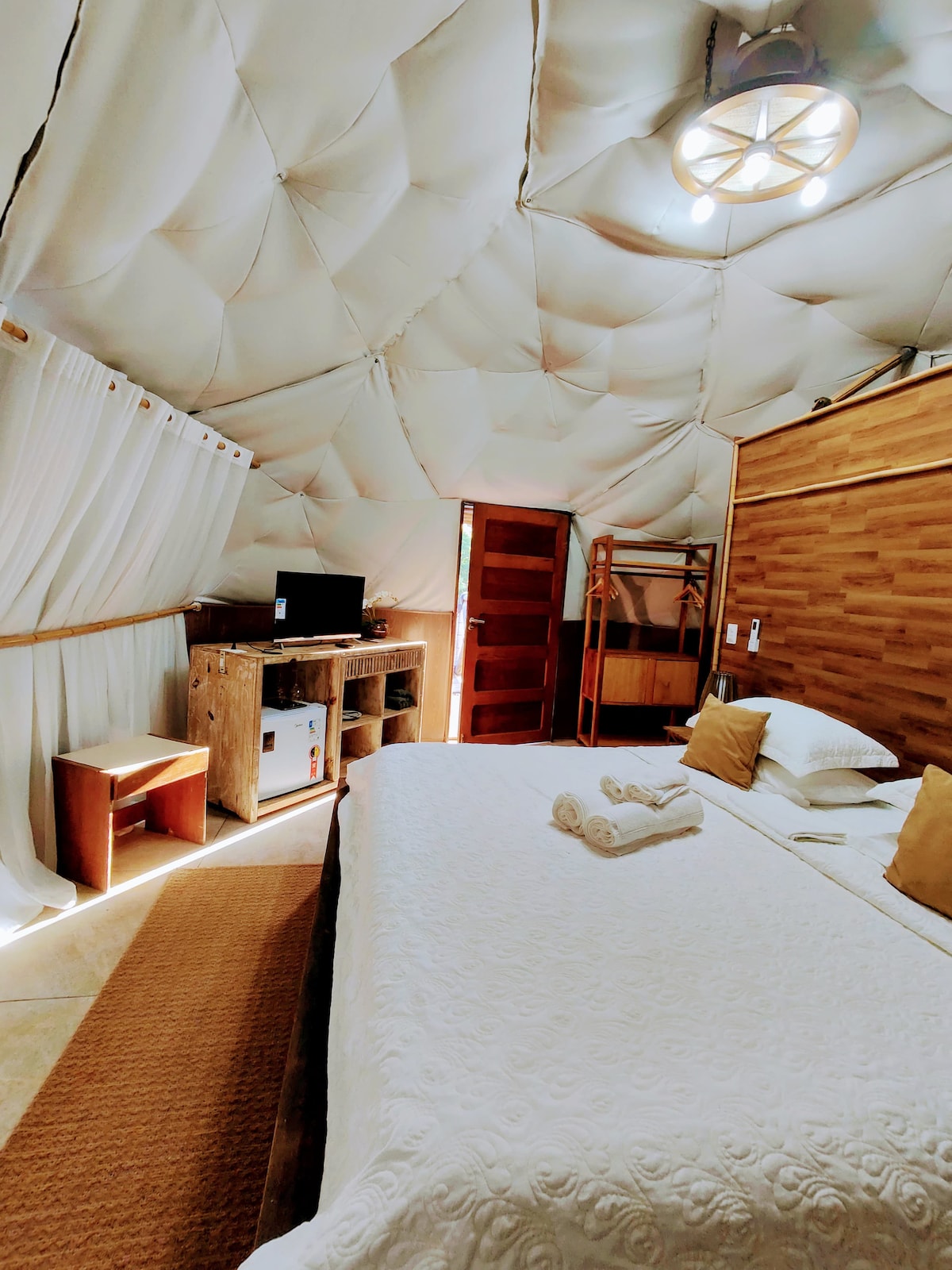 The unique glamping suite features a spacious bed adorned with neutral bedding and two accent pillows. Natural light fills the room through a small window, and wooden furnishings include a mini-fridge and a TV. Soft curtains frame the entrance, enhancing the cozy ambiance.