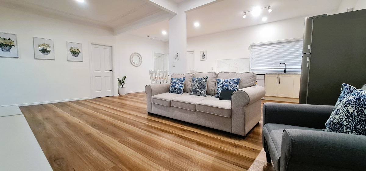 A modern living area features light-colored walls and hardwood flooring. A comfortable sofa with decorative cushions faces a spacious kitchen area, which is visible in the background. Natural light illuminates the space through windows and complements the minimalistic decor.
