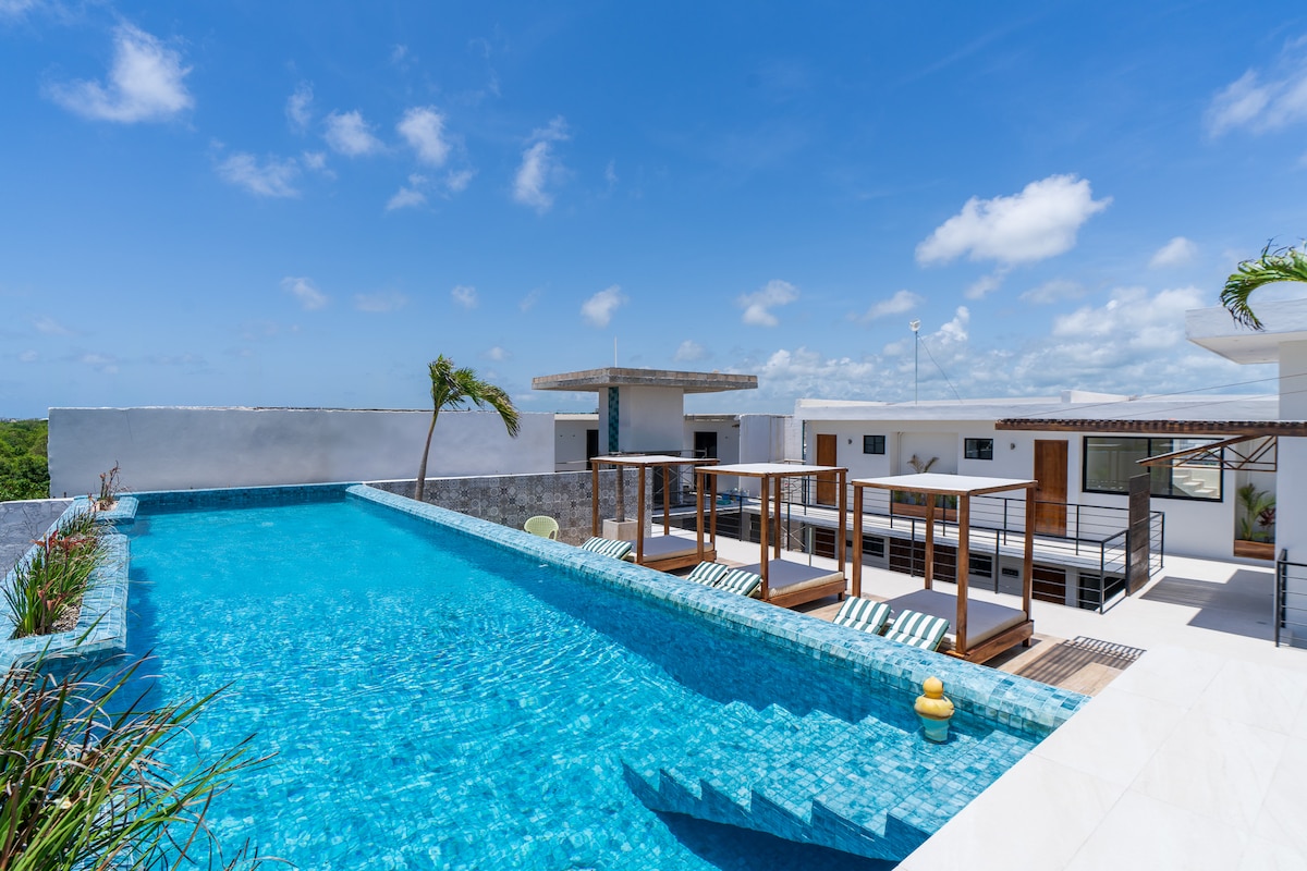 A rooftop pool is surrounded by modern lounge areas, offering a vibrant turquoise hue that contrasts with the clear blue sky. Sun loungers sit along the pool's edge, providing comfortable seating options for relaxation and leisure.