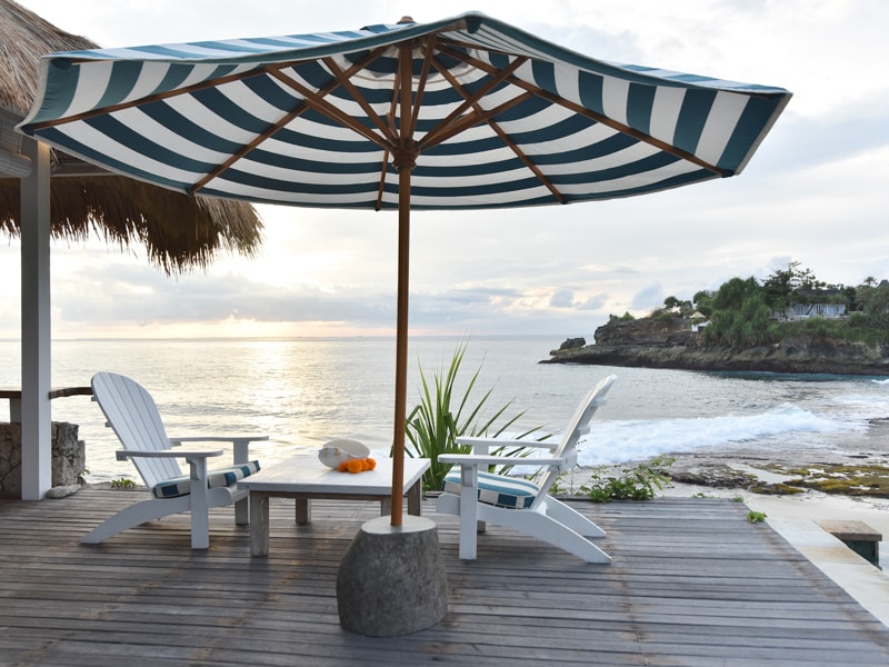 A beachside terrace features two white wooden chairs and a small table beneath a blue and white striped umbrella. The ocean can be seen in the background, with waves gently lapping at the rocky shore. The sky exhibits soft hues of orange and blue, indicating sunset.