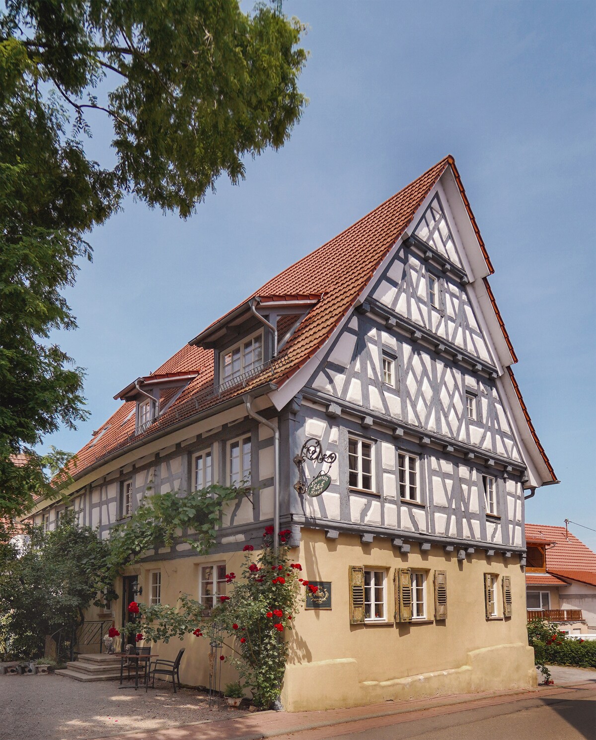 A charming historical timber-framed building features a sloped roof and decorative eaves. Lush greenery and vibrant red roses frame the entrance, while small outdoor seating is positioned against the façade. The clear blue sky provides a serene backdrop for this centrally located accommodation.