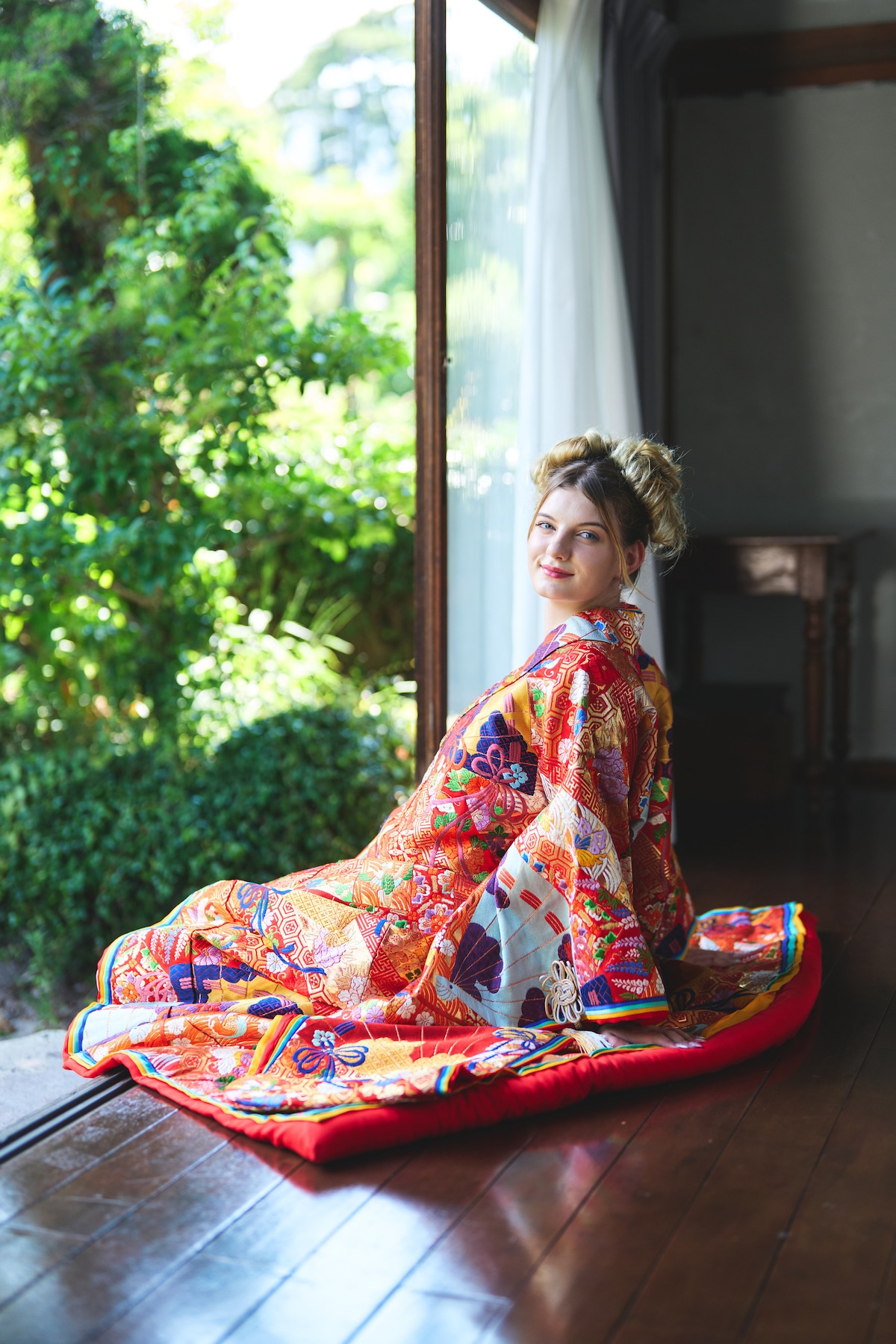 A person is seated gracefully on a wooden floor, wearing a vibrant traditional kimono adorned with intricate patterns. Natural light filters through a large window, illuminating the colorful fabric. Green foliage is visible outside, creating a serene and inviting atmosphere.