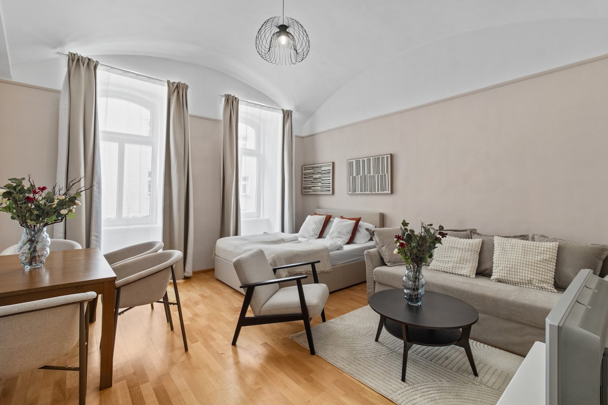 A well-lit studio features a spacious bed with crisp linens, a sofa, and two armchairs arranged around a round coffee table. A dining table is set for four. Natural light filters through large windows adorned with curtains, complementing the neutral color palette.