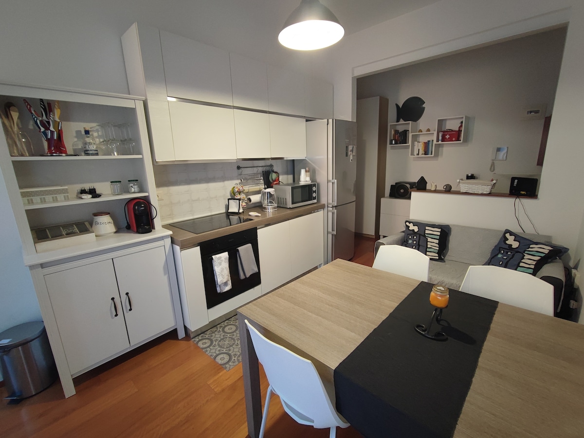 A modern kitchen area features sleek white cabinetry and a stylish countertop. Essential appliances, including a coffee maker and microwave, are visibly arranged. A dining area is set with a simple table and white chairs, complemented by decorative cushions on the nearby sofa.
