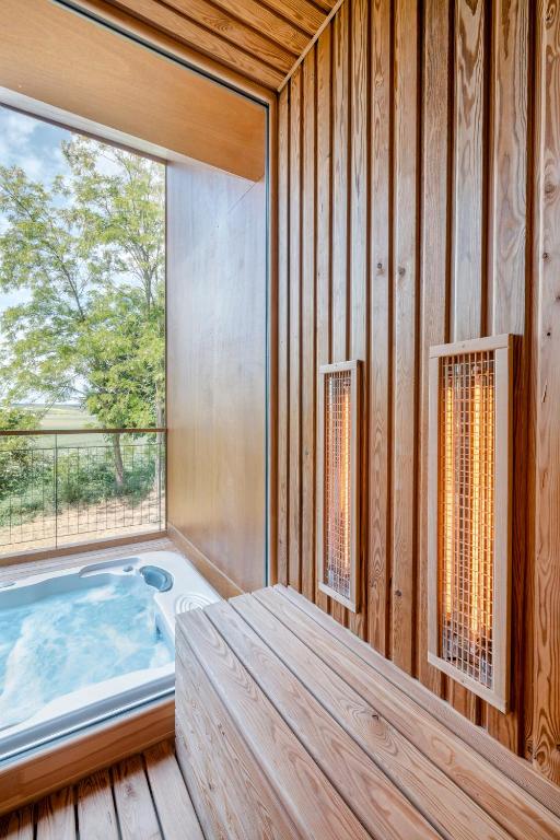 A serene space showcases an inviting hot tub next to a large window, allowing natural light to illuminate the wooden walls and bench. Infrared heaters are installed on the walls, and a lush green landscape is visible through the window.