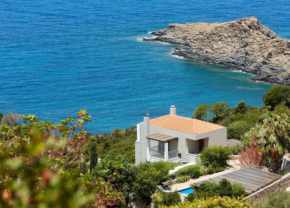 A modern, two-story home is nestled among lush greenery, with a terracotta roof complementing the serene landscape. The property overlooks the blue sea, where rocky outcrops emerge from the water, creating a peaceful coastal scene.