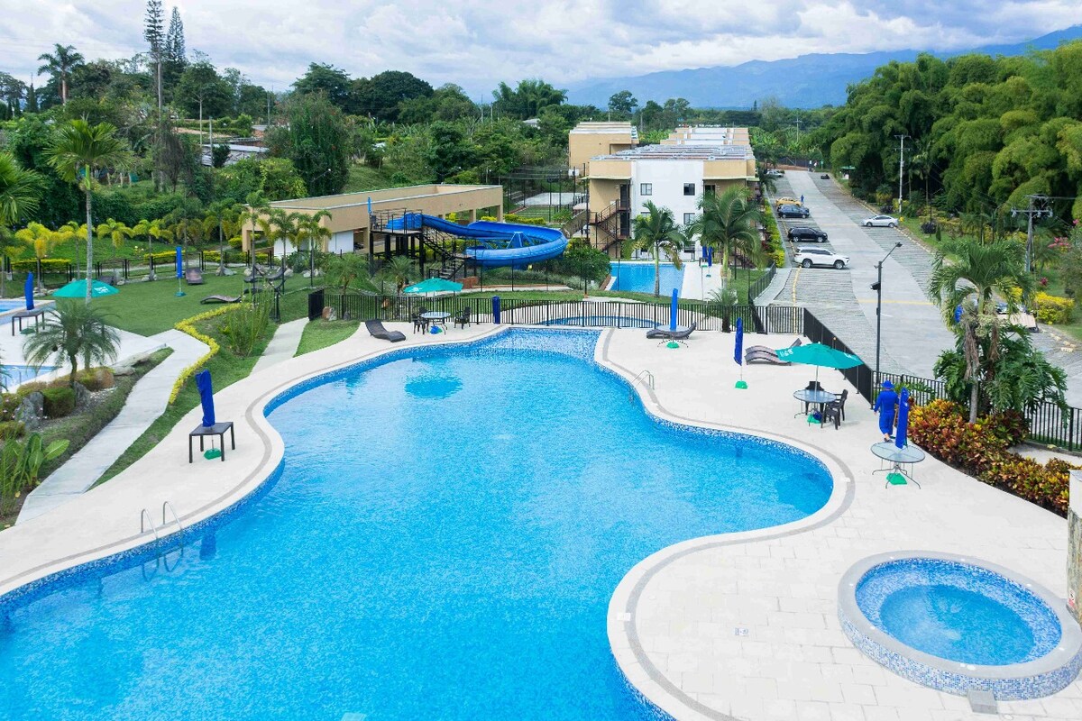 A large outdoor swimming pool features a unique shape, surrounded by palm trees and sun loungers. A smaller jacuzzi is visible nearby, along with a water slide. Lush greenery and a manicured lawn are seen in the background, offering a tranquil setting.