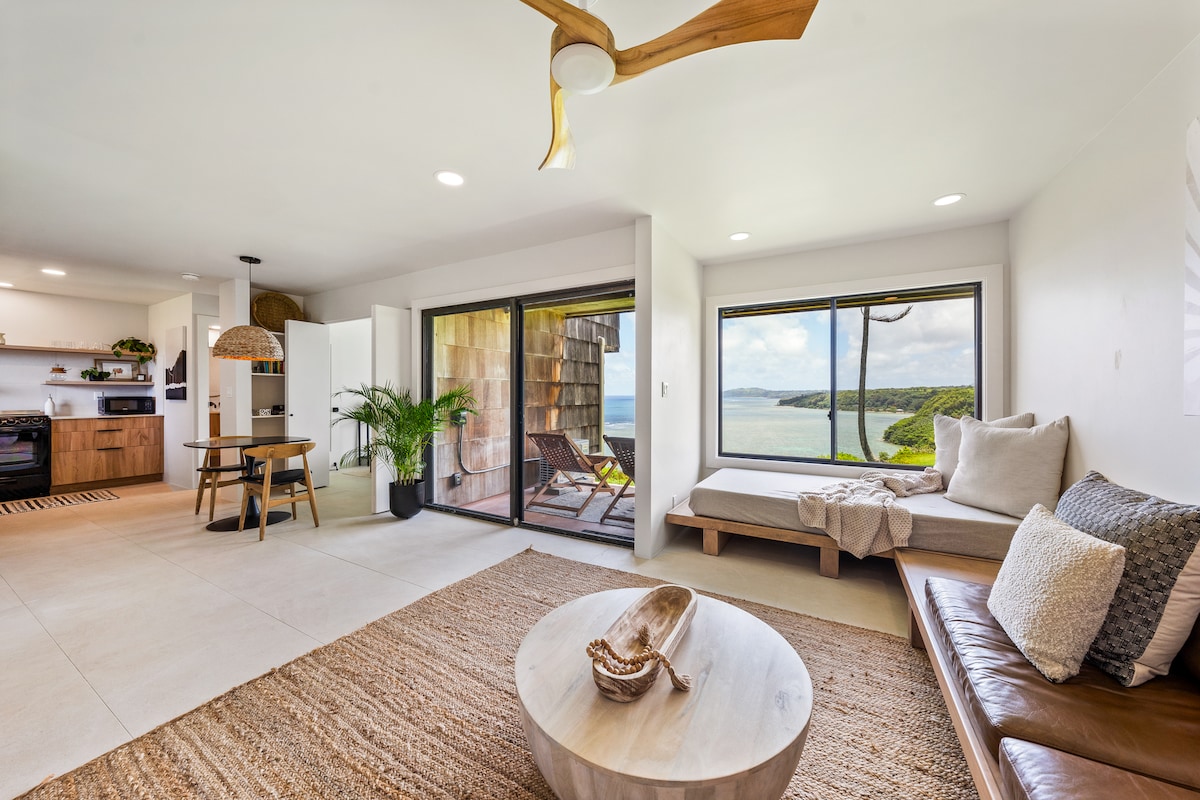A spacious living area features large glass doors that open to a beautiful ocean view. A cozy window seat is positioned beside the view, while a round natural fiber rug lies centrally on the floor. The modern kitchen is visible in the background, complemented by warm wood finishes.