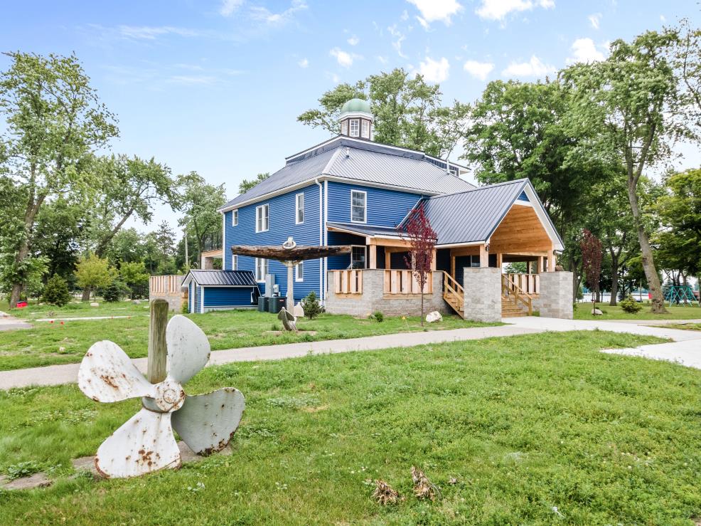 An exterior view of a newly renovated blue building with a distinctive rooftop feature. Surrounded by green grass and trees, this structure includes a deck area with outdoor seating and is complemented by a large decorative propeller sculpture in the foreground.