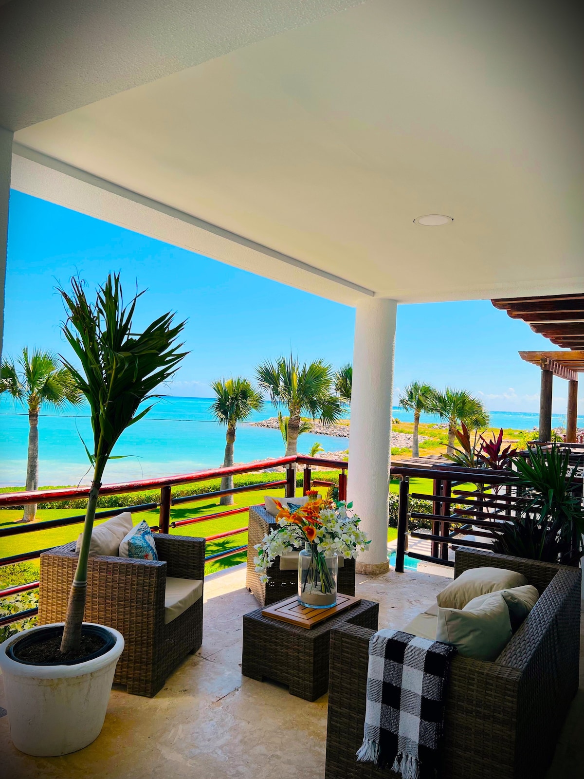 A private terrace features comfortable seating with woven furniture and vibrant cushions. A potted palm adds a touch of greenery. The view showcases the expansive Caribbean Sea, framed by swaying palm trees and a clear blue sky, providing a serene outdoor space for relaxation.