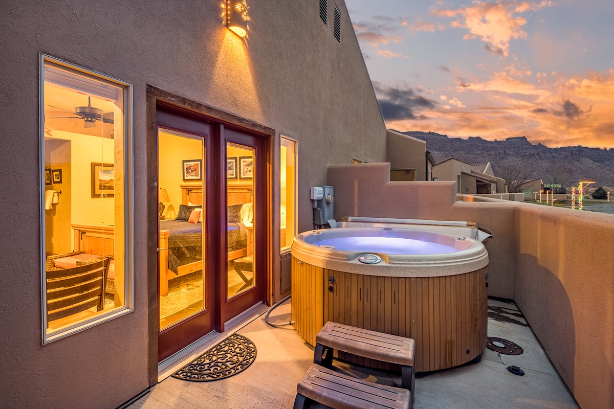 An inviting outdoor patio features a private hot tub surrounded by a low wall. Glass doors lead into the interior, showcasing warm light from inside. Mountain views are visible in the background, with colors of sunset casting a serene ambiance.