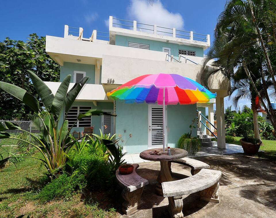 A spacious outdoor area features a large, multicolored umbrella over a circular stone table, surrounded by tropical greenery. The building showcases a light blue exterior with multiple windows, and a second-floor patio is visible above the entrance.