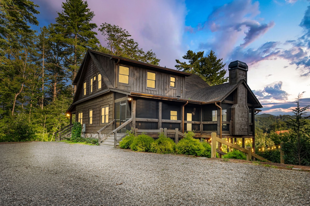 A modern cabin is set against a backdrop of pine trees, with a gravel driveway leading to a welcoming exterior. Large windows reflect the natural light and surroundings, while a screened porch offers a glimpse of outdoor living spaces, inviting relaxation in the wilderness.