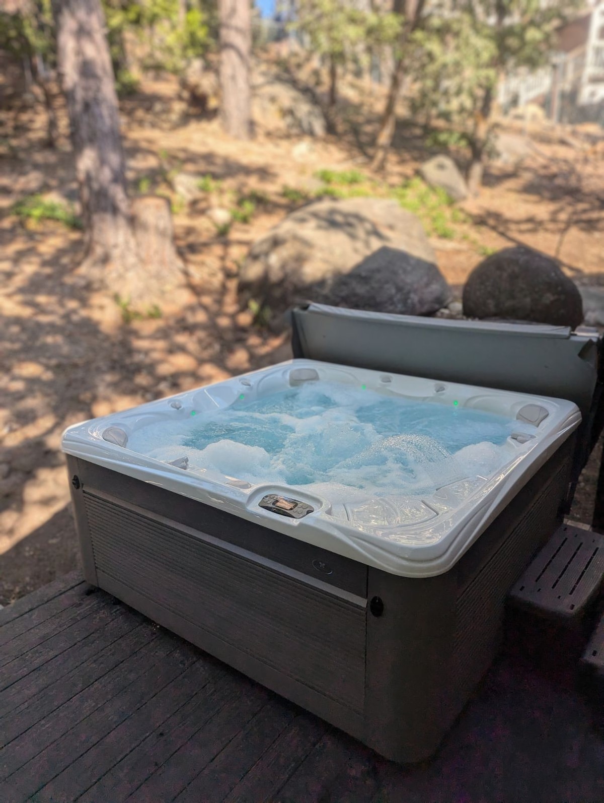 A hot tub is positioned on a wooden deck, surrounded by natural elements such as trees and rocks. Bubbles rise from the water's surface, indicating a warm, inviting setting for relaxation.