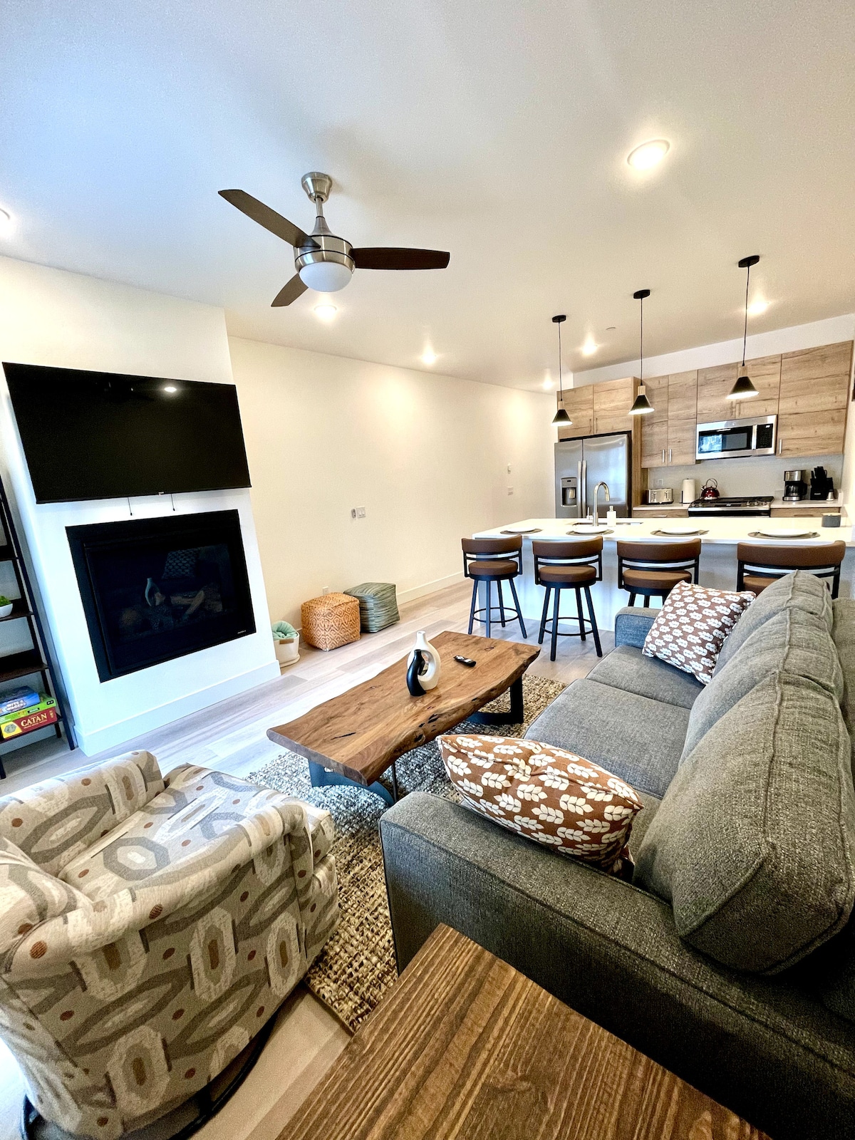 A modern living area is presented, featuring a cozy gray sofa with patterned cushions and a wooden coffee table. A ceiling fan and wall-mounted TV add functionality. An open kitchen with bar seating can be glimpsed in the background, alongside warm wooden accents and softly lit fixtures.