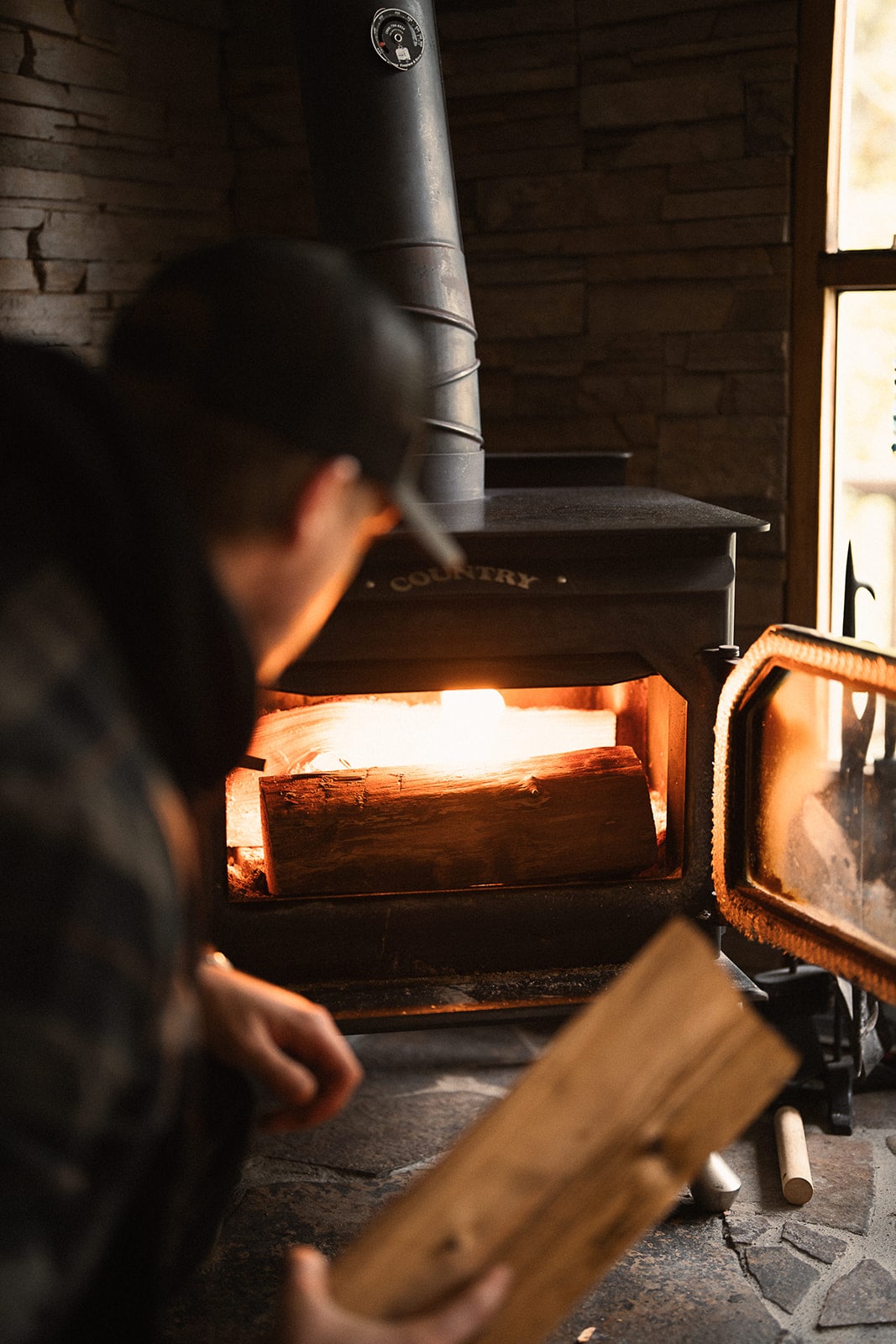 A woodstove is positioned in the corner of a cozy cabin, showcasing a flickering flame within. Logs are stacked inside, with a partially open glass door revealing warm light. The stone wall behind the stove adds a rustic touch to the inviting atmosphere.