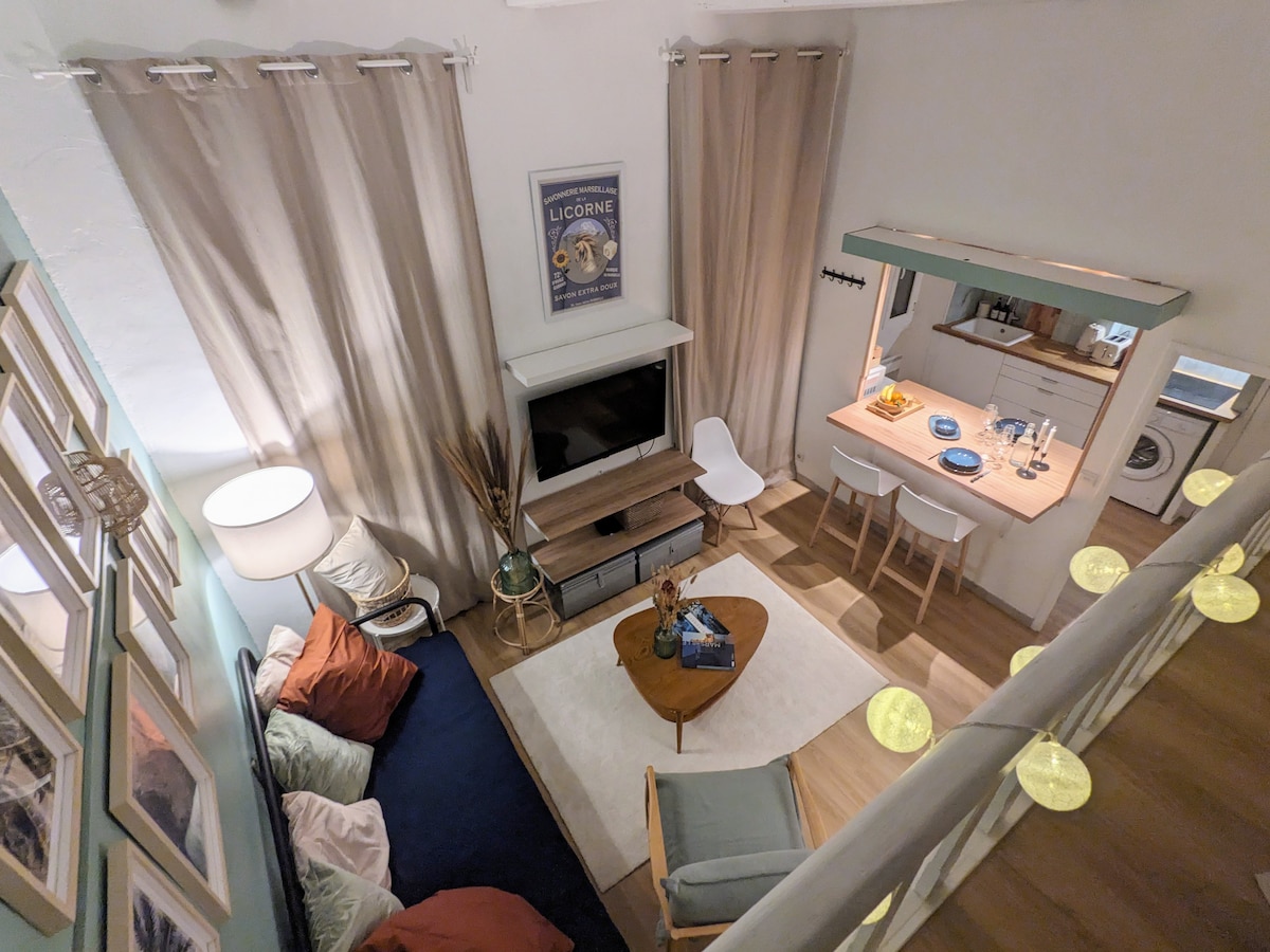 The living area features a cozy seating arrangement with mixed cushions on a deep blue rug. A wooden coffee table sits at the center, accompanied by a simple dining setup in the adjacent kitchenette. Curtains frame the windows, and a flat-screen TV is mounted on the wall.