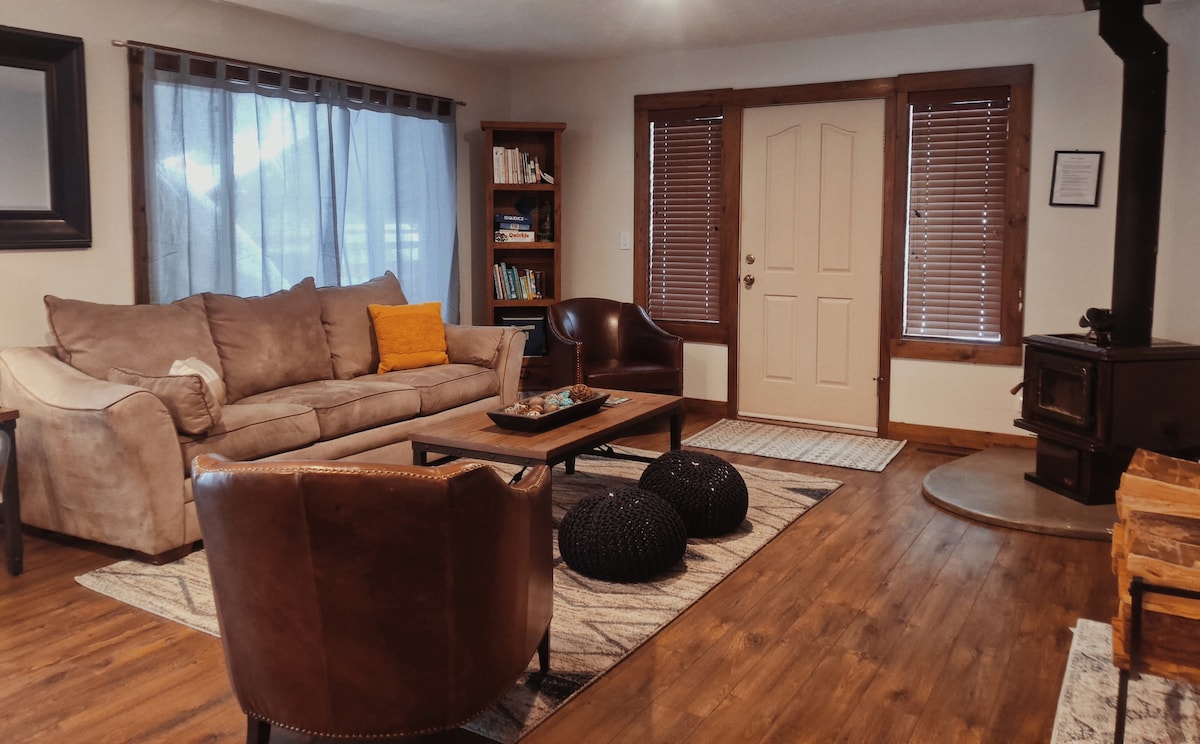 A welcoming living room is highlighted by a large, comfortable sofa and two armchairs. A wooden coffee table sits at the center, surrounded by decorative poufs. Natural light enters through the sheer window curtains, and a quaint wood stove adds warmth to the space.