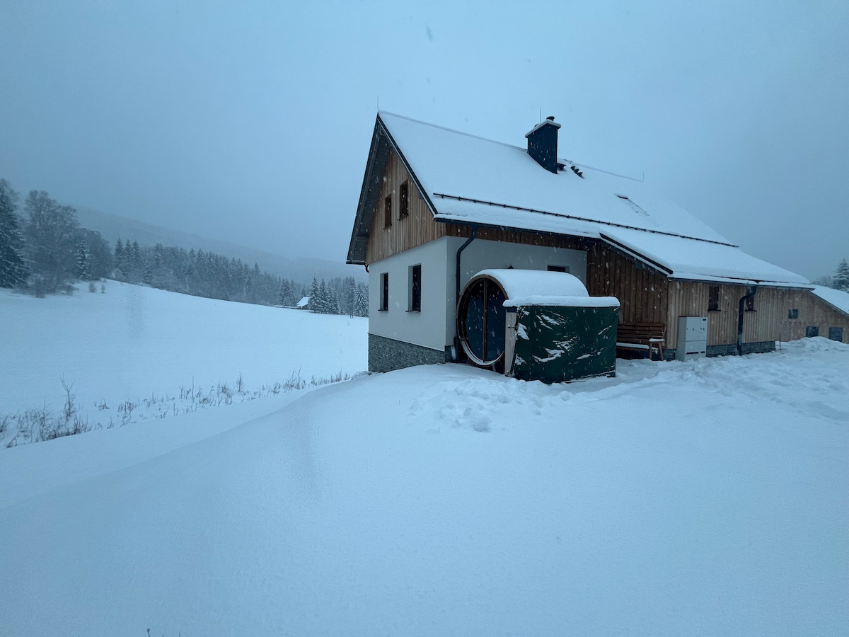 A wooden chalet is seen covered in fresh snow, set against a serene winter landscape. The structure features a sloped roof and a small attached shed. Snow blankets the ground, creating a tranquil scene amidst the surrounding fields and trees.