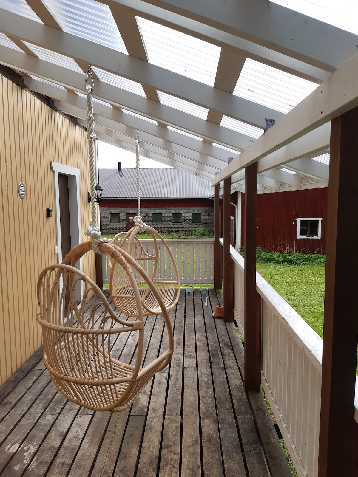 Two woven hanging chairs are suspended from the ceiling on a covered wooden terrace. The space is lined with white railings, while the wooden floor provides a natural touch. A grassy area is visible beyond the railing, enhancing the outdoor ambiance.