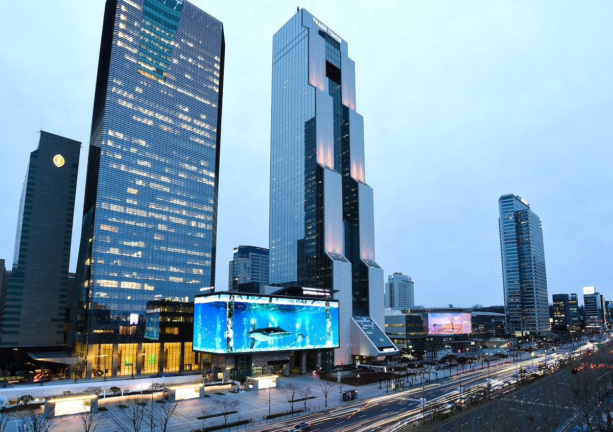 A modern city skyline is displayed, featuring tall skyscrapers with illuminated facades. A large digital screen showcasing vibrant images is positioned prominently at the base of one building. The scene captures a dynamic urban environment during dusk, with a wide thoroughfare visible in the foreground.