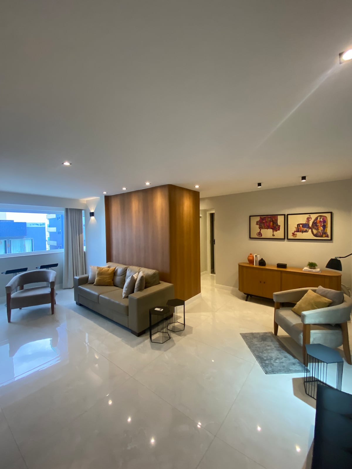 A spacious living area features a neutral palette, with a comfortable sofa and two armchairs arranged around a low table. Light reflects off the polished tile floor, enhancing the room's brightness. Artwork adorns the wall, and large windows allow natural light to fill the space.