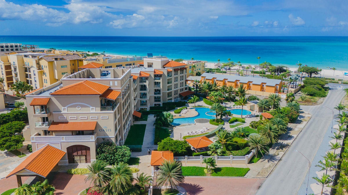 An aerial view captures the luxurious condominium surrounded by lush palm trees and well-maintained gardens. The clear blue pool is visible in the courtyard, with the pristine beach and calm waters of Eagle Beach in the background, adding to the serene atmosphere.