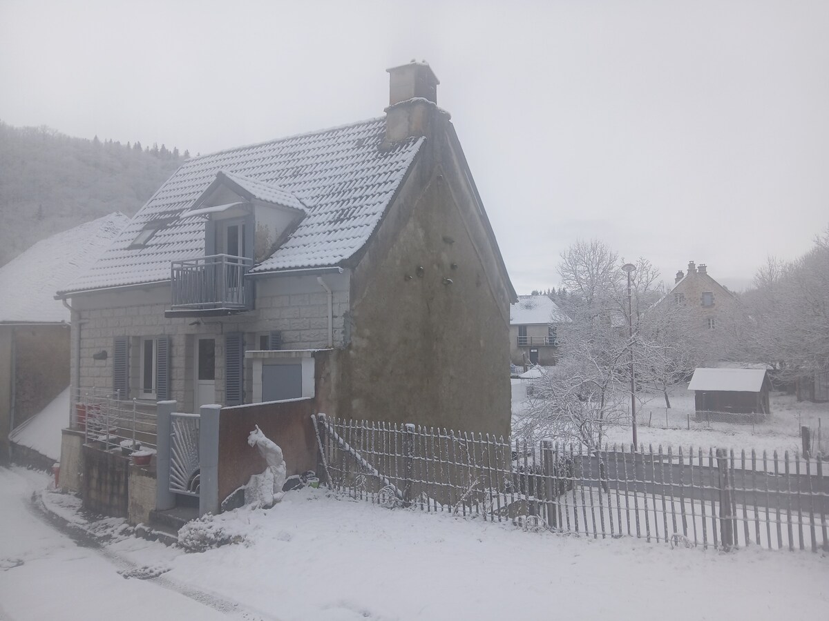 A quaint two-story house is seen blanketed in fresh snow, with a sloped roof and a small balcony. Surrounding trees and a fence are coated in white, providing a serene winter landscape. Nearby structures and snow-covered ground contribute to the tranquil ambiance.