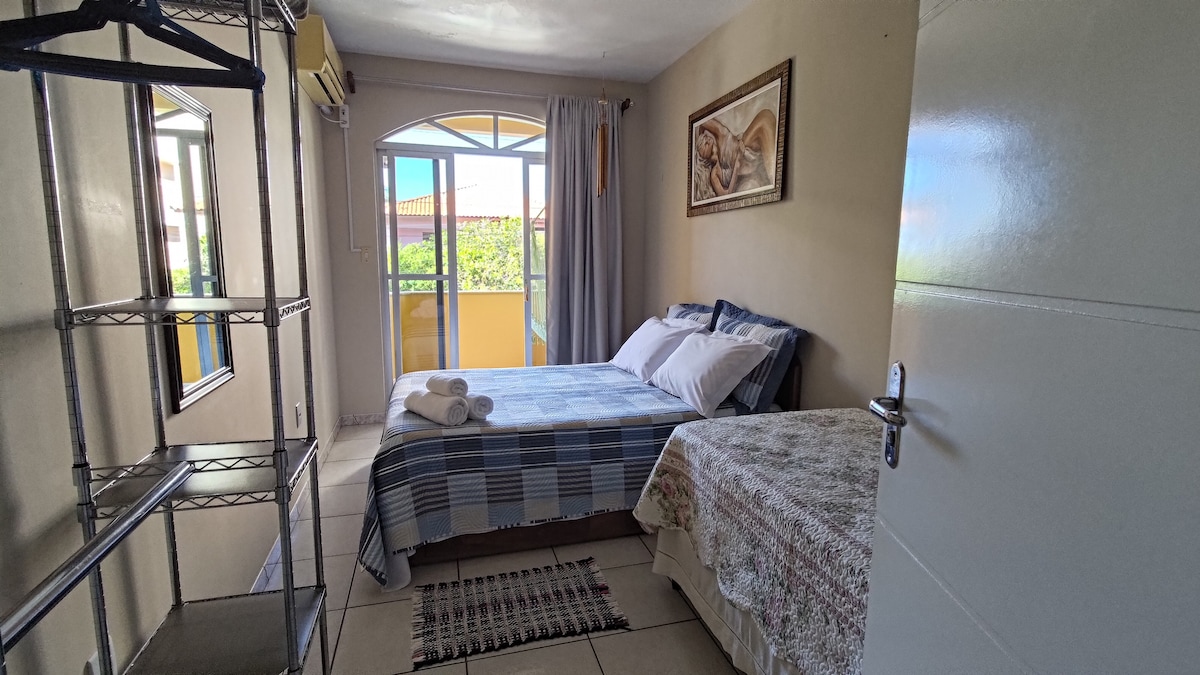 A well-lit bedroom is showcased, featuring a spacious bed with neatly arranged bedding. A second bed is positioned nearby. Natural light filters through a large window adorned with light curtains, leading to a small balcony. A shelving unit is visible for storage, enhancing functionality.