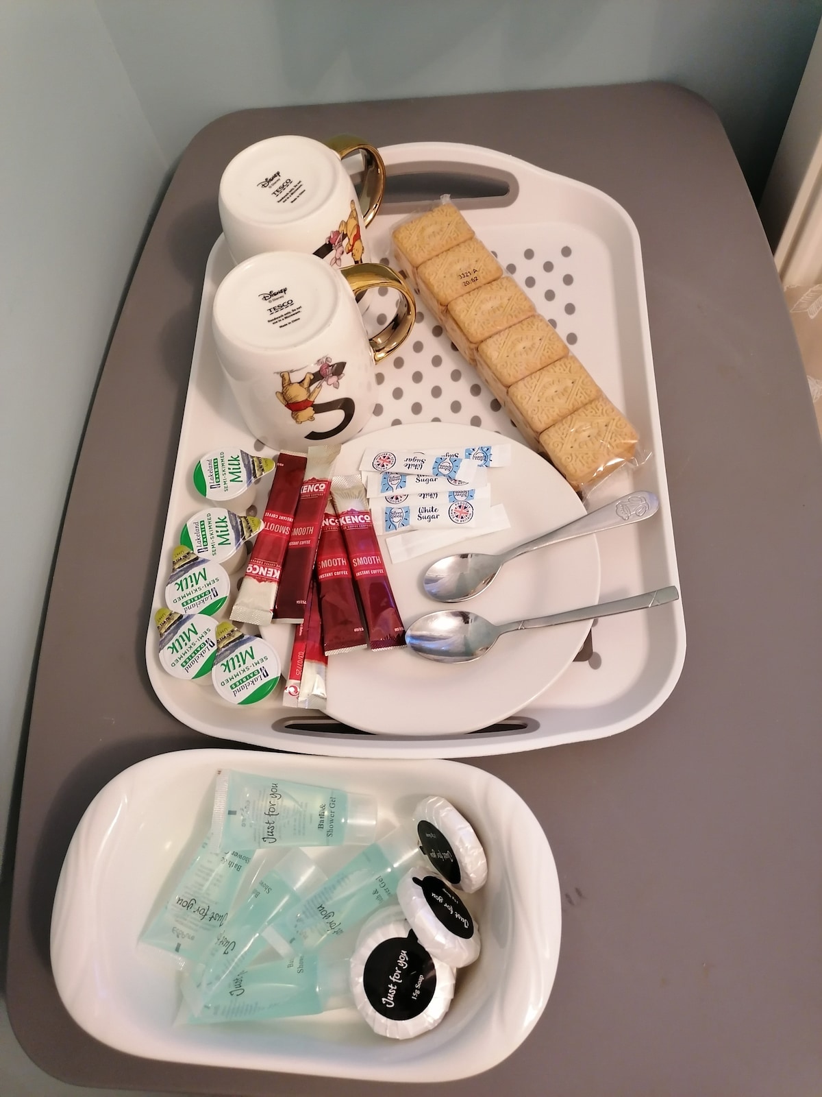 A welcoming tray is displayed with two mugs, packets of tea and coffee, and several sugar sachets arranged neatly. Silver spoons rest beside a stack of biscuits. A bowl holds complimentary toiletries, including miniature bottles and soap bars, providing essentials for guests.