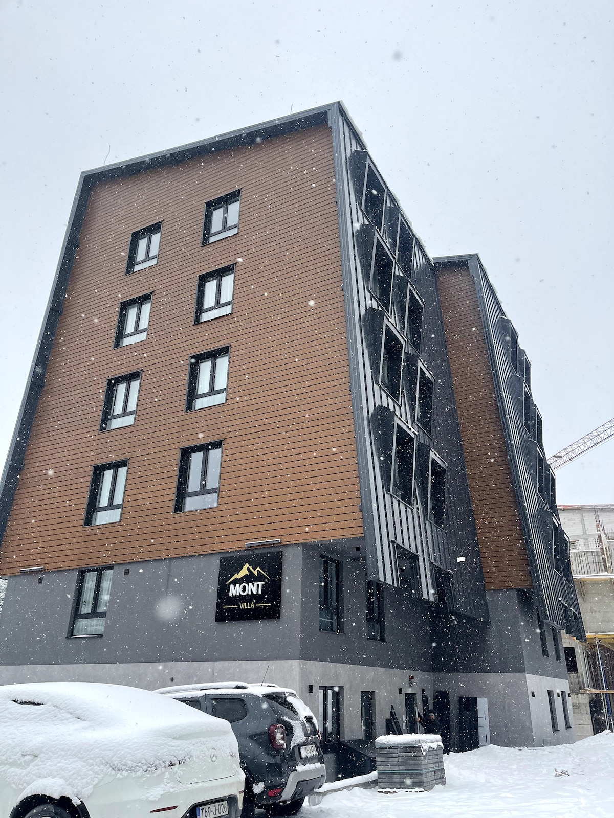 A modern building features a combination of dark and wooden facade elements. Snow is visibly covering the ground and car parked in front. Multiple windows are displayed, allowing for natural light, while a sign reading 'MONT' is mounted at the base.
